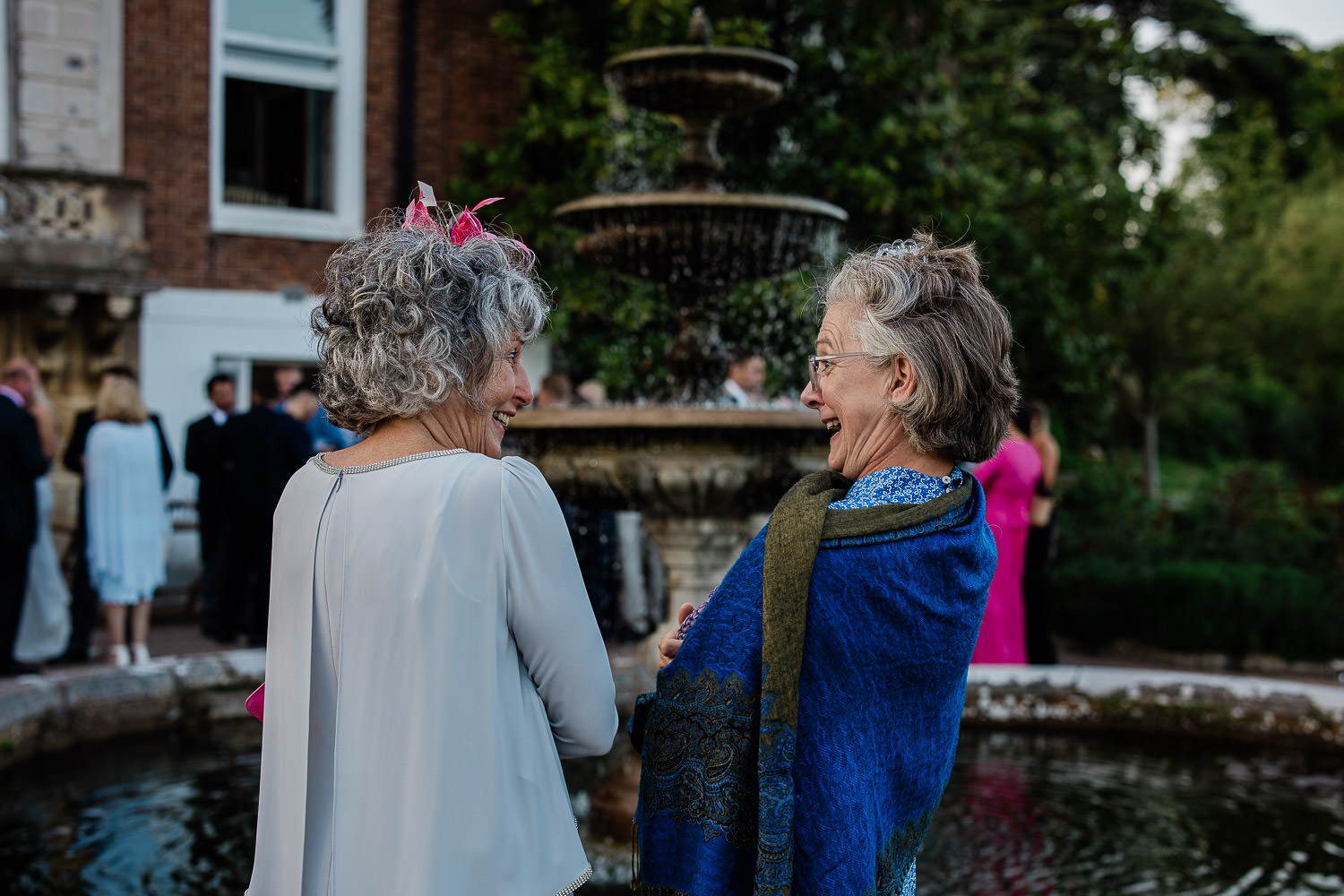 A candid, reportage moment between two women who share a laugh together, during an outdoor wedding reception in a rose terrace.