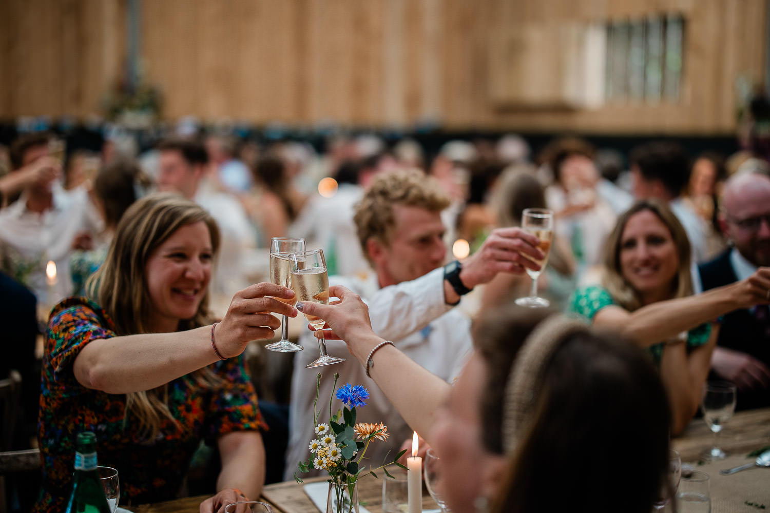 Guests share a cheers at the end of a wedding speech, during a reception in a boho inspired English barn in Devon.