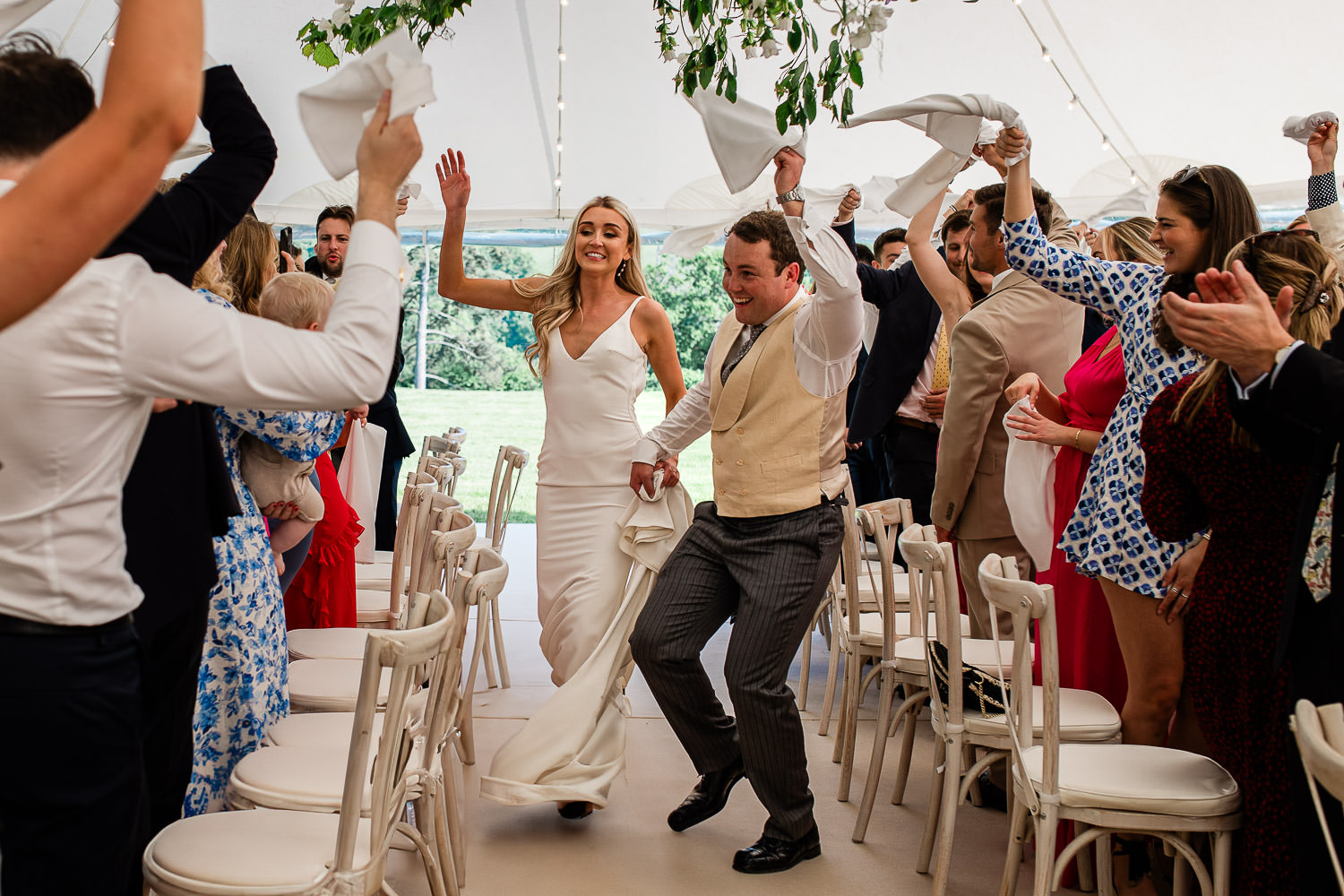 A bride and groom run down the aisle between tables during a marquee wedding reception, while wedding guests wave round napkins to welcome them.