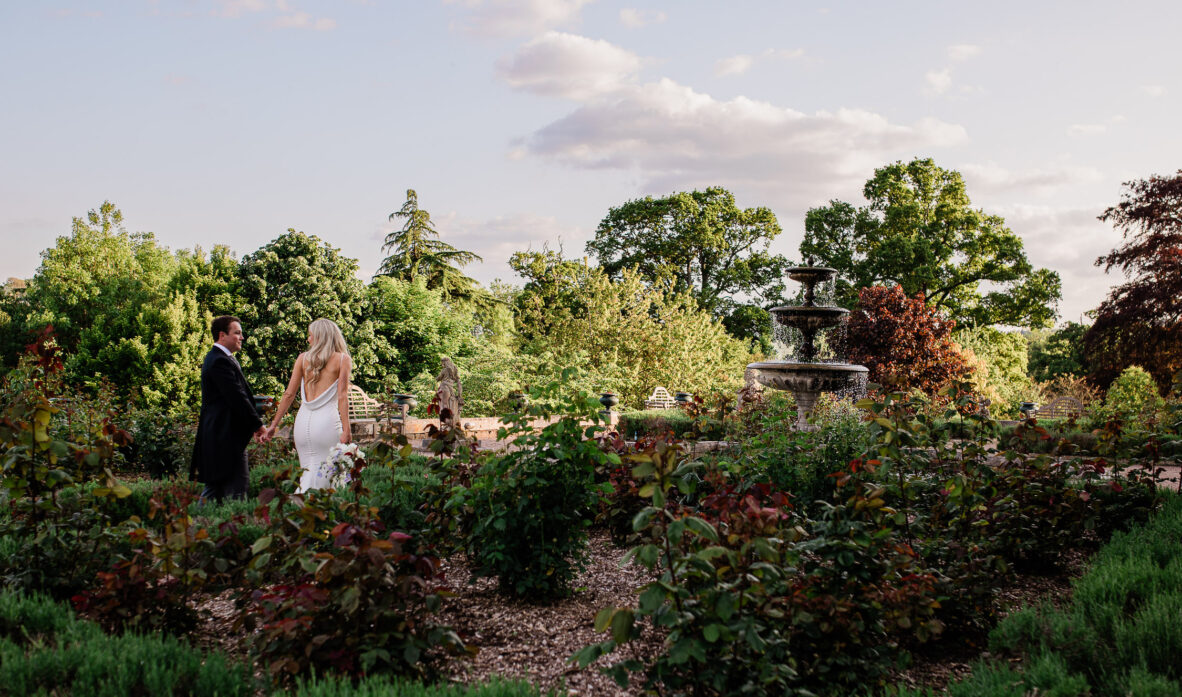 A newly wed couple walk through the rose terrace of an english country house wedding venue, during documentary style wedding portraits.