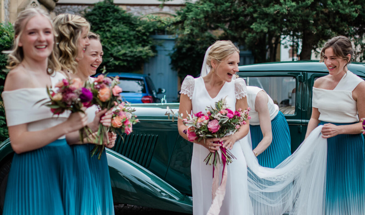 A bride steps out of a vintage car and looks excitedly at her friends, before entering the religious ceremony at Topsham Methodist Church.