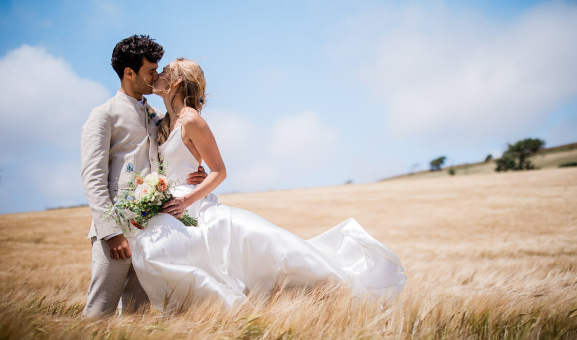 A wedding couple share  a kiss in a barley field, during a quiet moment away from their outdoor bohemian inspired wedding in the UK.